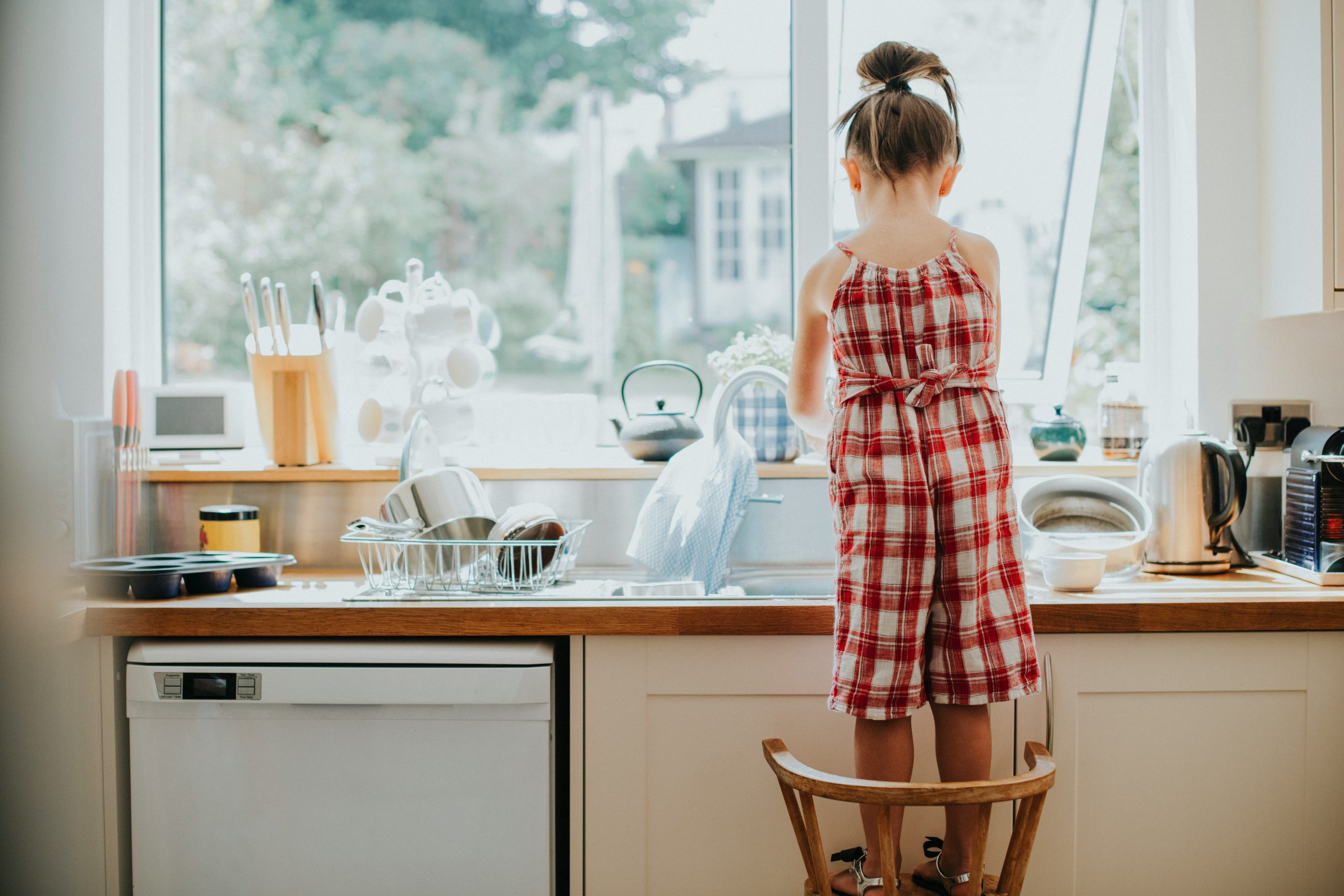 Little girl standing on a chair to wash dishes at the sink, enjoying a safe and healthy routine made better with Brita Pro filtered water.