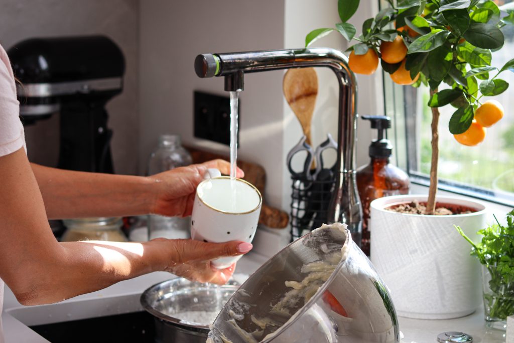 Close-up of a woman rinsing a ceramic mug under a running kitchen faucet beside a sunlit window, highlighting the clarity and safety of Brita Pro filtered water in a modern, eco-conscious home.