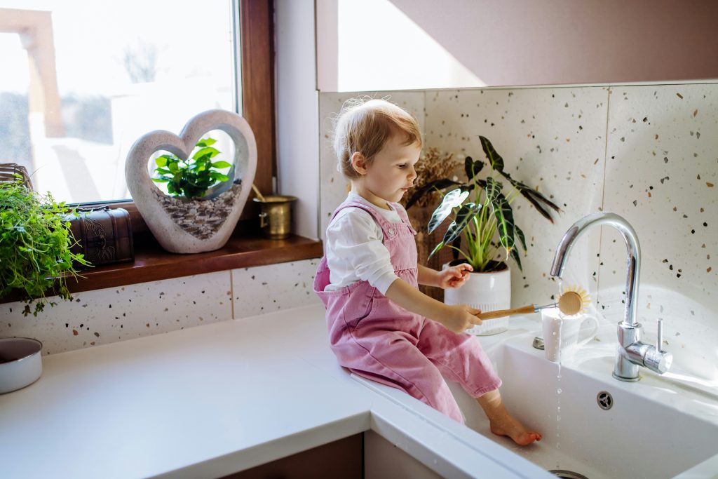 Toddler girl playing at a kitchen sink with a dish brush and running clean water filtered by Brita Pro at home.