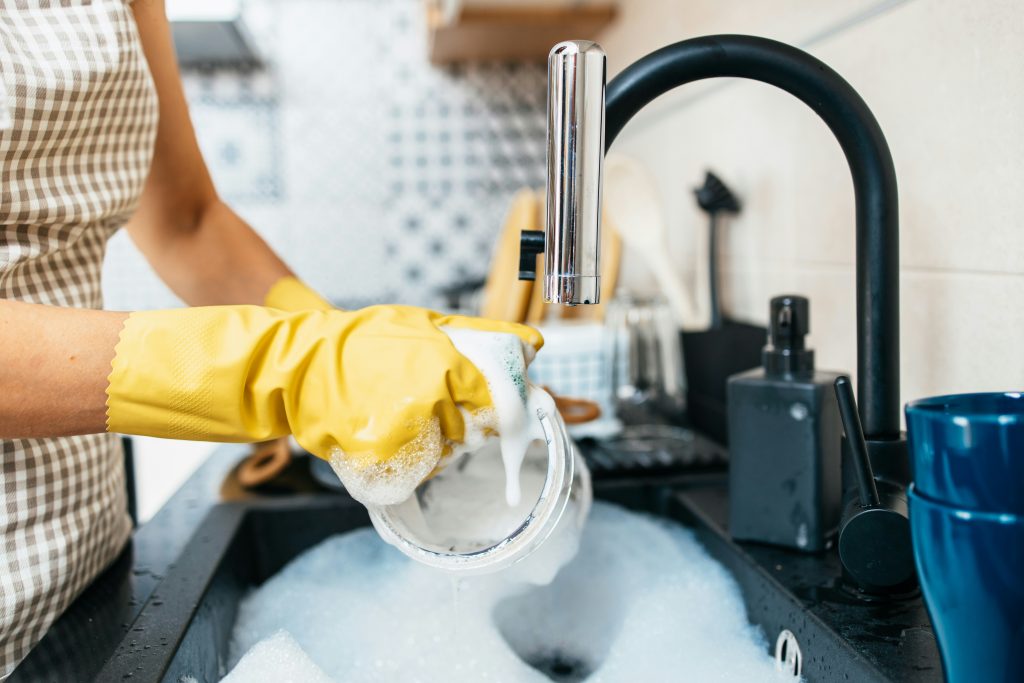 Close-up of a person wearing yellow gloves washing a soapy glass under a modern black kitchen faucet, promoting home hygiene and the benefits of clean, filtered water with Brita Pro water filtration systems.