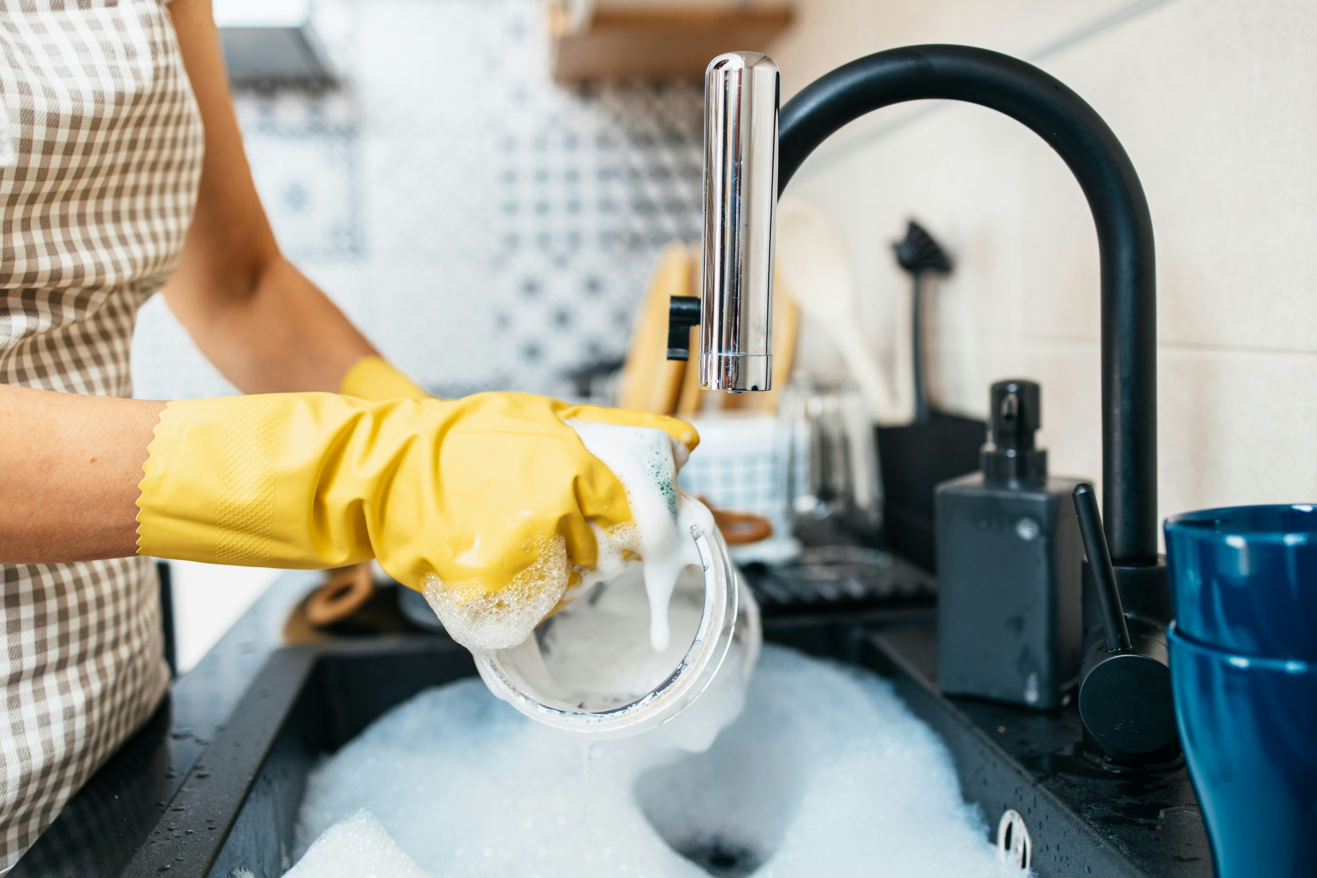 Close-up of a person wearing yellow gloves washing a soapy glass under a modern black kitchen faucet, promoting home hygiene and the benefits of clean, filtered water with Brita Pro water filtration systems.