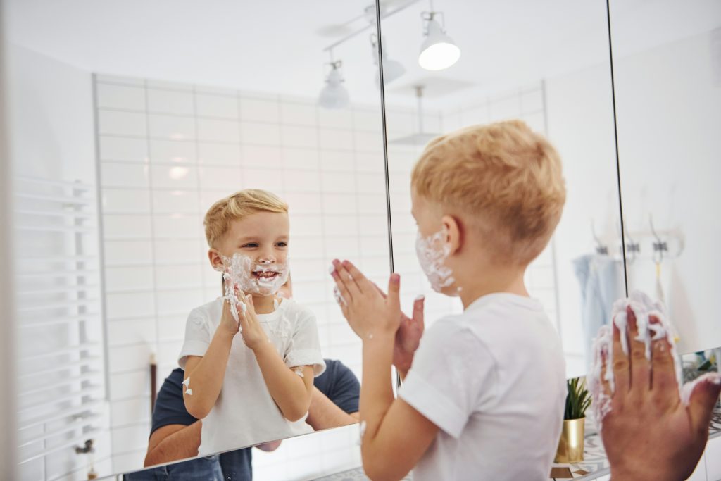 Young boy playfully pretending to shave with shaving cream in front of a bathroom mirror, guided by a parent using filtered water from Brita Pro