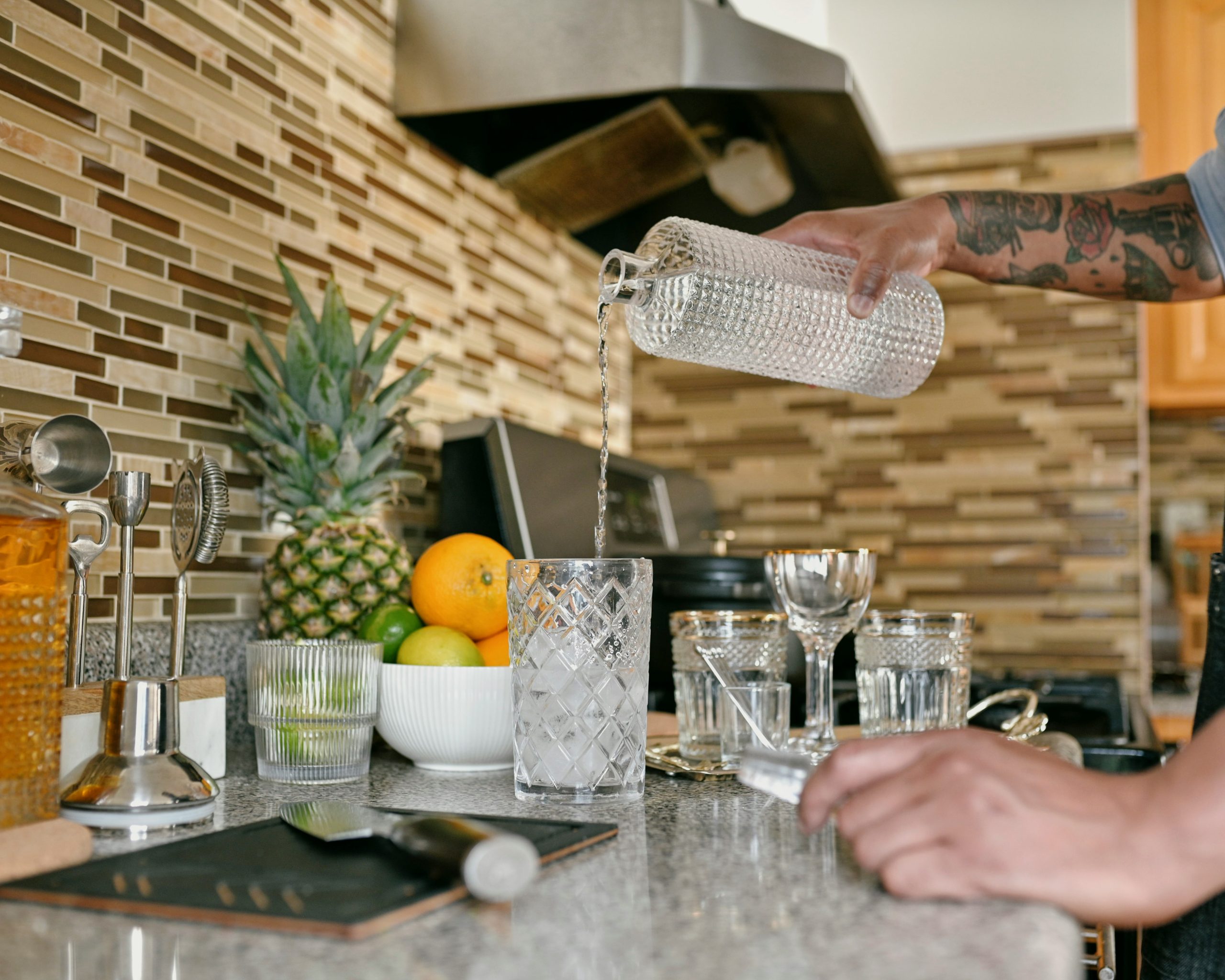 Tattooed hand pouring crystal-clear water from a glass pitcher into an ice-filled tumbler on a modern kitchen counter, showcasing healthy hydration and the purity delivered by Brita Pro whole-home water filtration systems