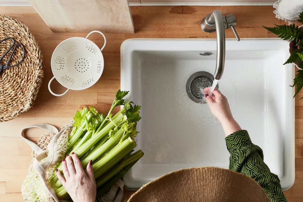 "Top-down view of a woman rinsing fresh celery under running tap water in a modern kitchen, highlighting healthy food prep, clean water, and eco-conscious living with Brita Pro water filtration.
