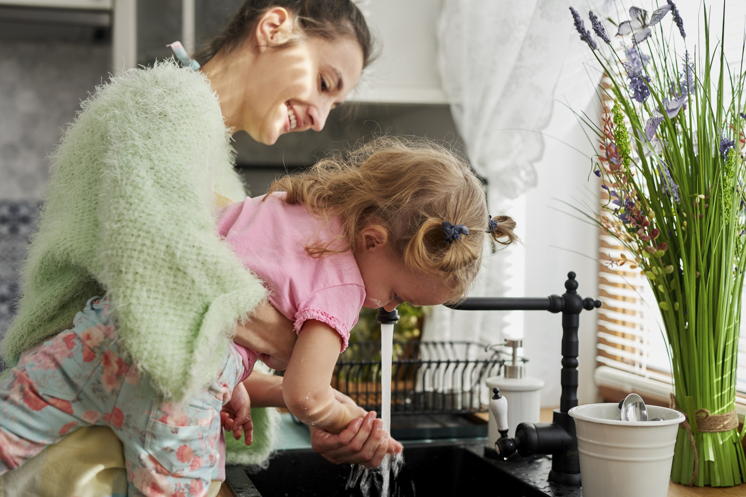 Smiling mother helping her toddler wash hands at the kitchen sink, highlighting the role of Brita Pro filtered water in promoting hygiene and safe routines in family-friendly homes.