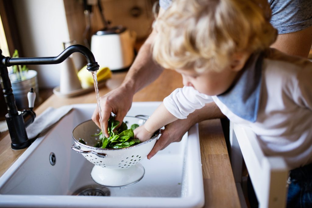 Young child helping wash fresh spinach with a parent under a running faucet—emphasizing the importance of clean, filtered water at home with Brita Pro’s whole-house water filtration system.
