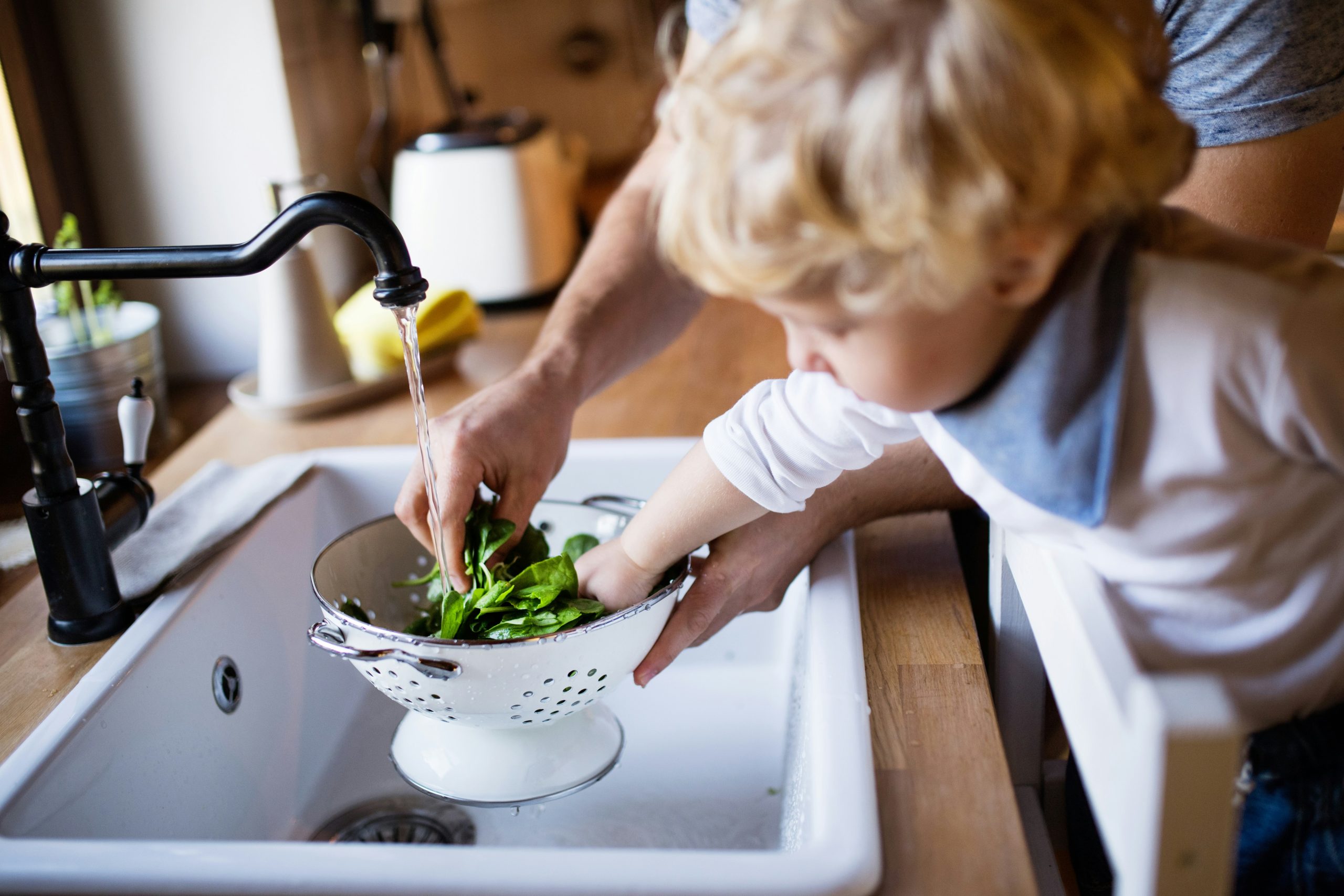 Young child helping wash fresh spinach with a parent under a running faucet—emphasizing the importance of clean, filtered water at home with Brita Pro’s whole-house water filtration system.