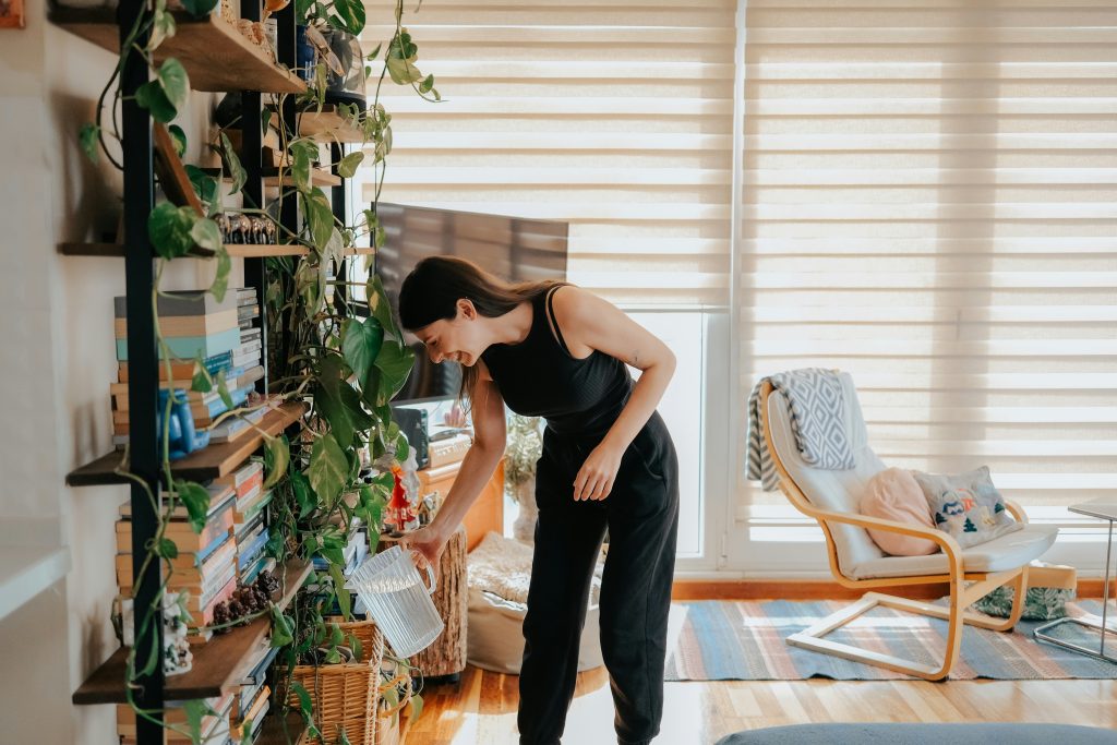 Smiling woman watering indoor plants with a clear pitcher in a cozy, sunlit living room—illustrating the everyday use of clean, filtered water from a Brita Pro home filtration system.