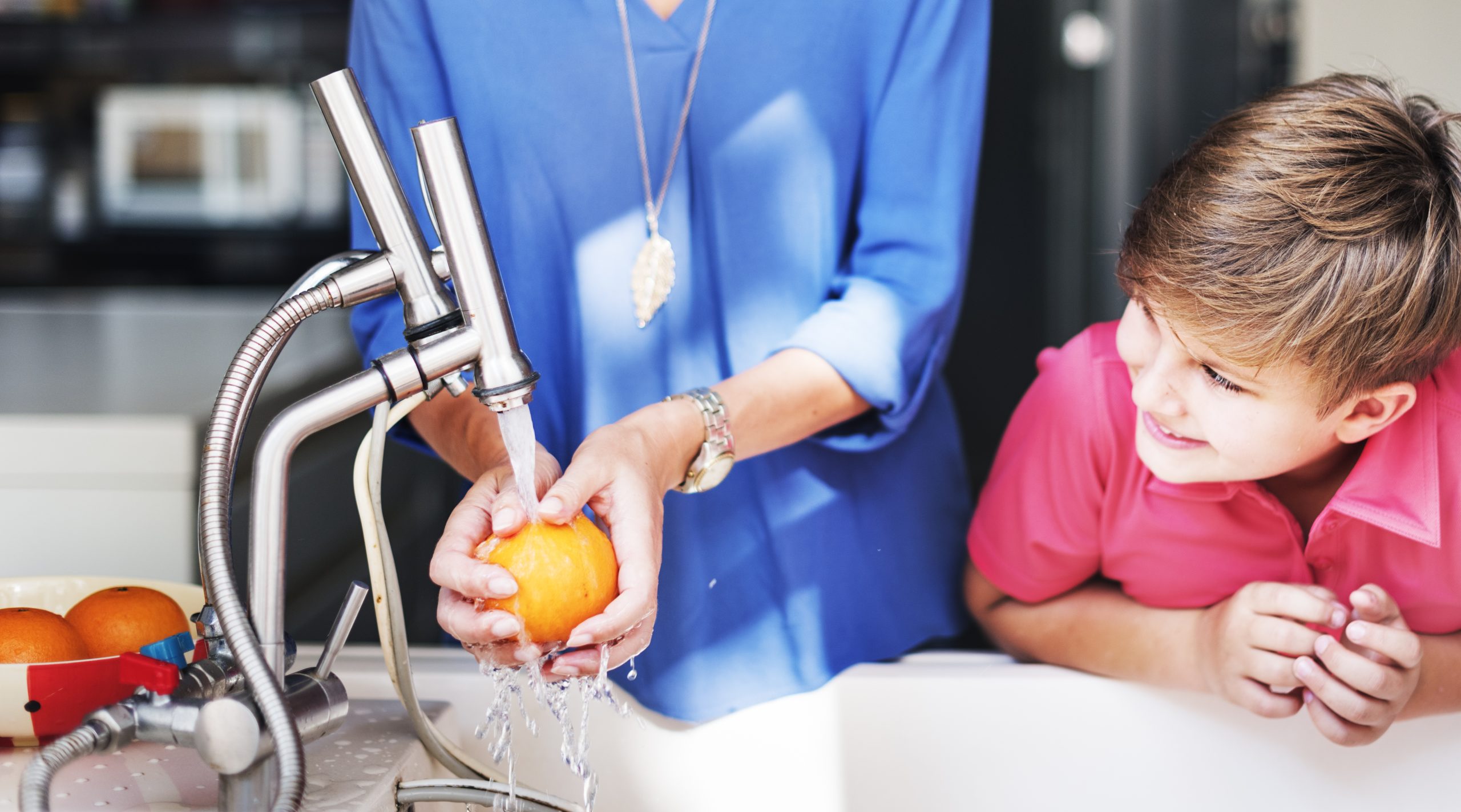 Smiling child watching as a parent washes a fresh orange under a modern kitchen faucet—promoting family wellness and the importance of Brita Pro’s water filtration for safe, clean produce.
