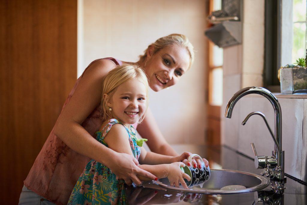 Smiling mother and daughter washing dishes together with filtered Brita Pro water at the kitchen sink, highlighting family bonding and clean water solutions.