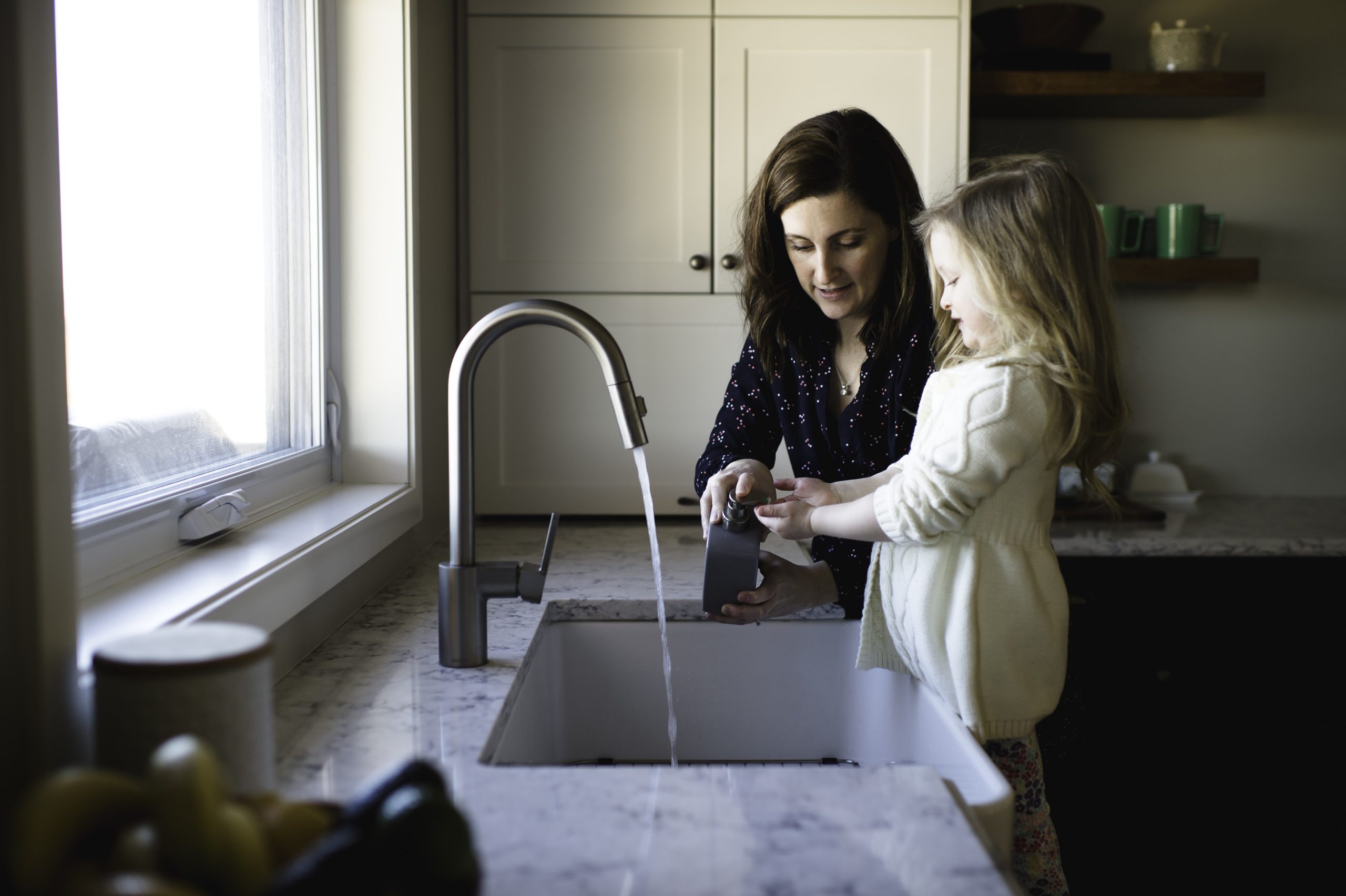 Mother helping daughter wash hands at a modern kitchen sink with running water, highlighting the everyday health benefits of Brita Pro’s whole-house water filtration system.